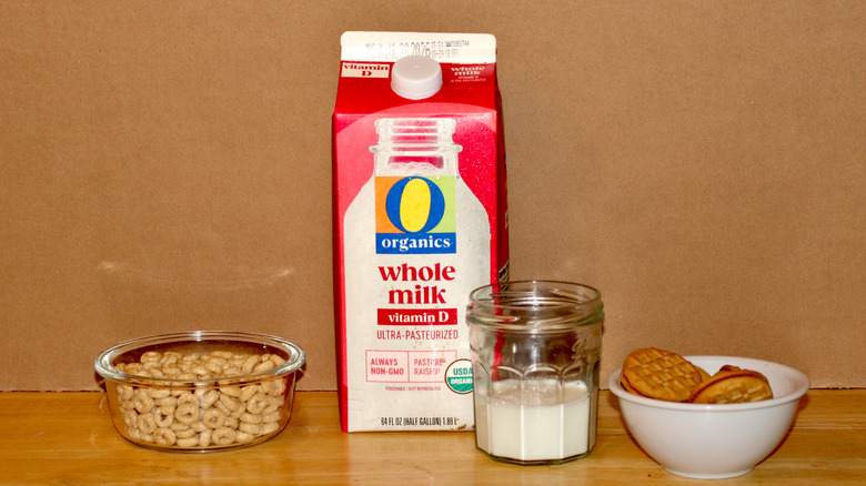 An O Organics brand carton of organic whole milk beside a glass of milk, a bowl of cereal, and some cookies