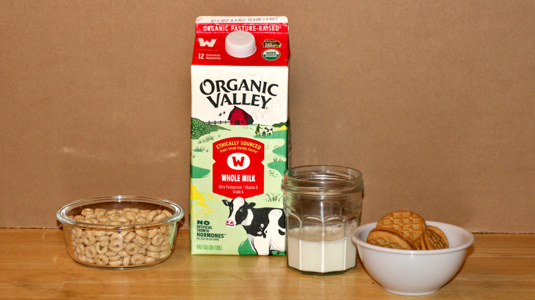 An Organic Valley brand carton of organic whole milk beside a glass of milk, a bowl of cereal, and some cookies