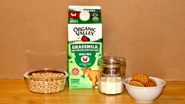 An Organic Valley brand carton of organic grass fed whole milk beside a glass of milk, a bowl of cereal, and some cookies