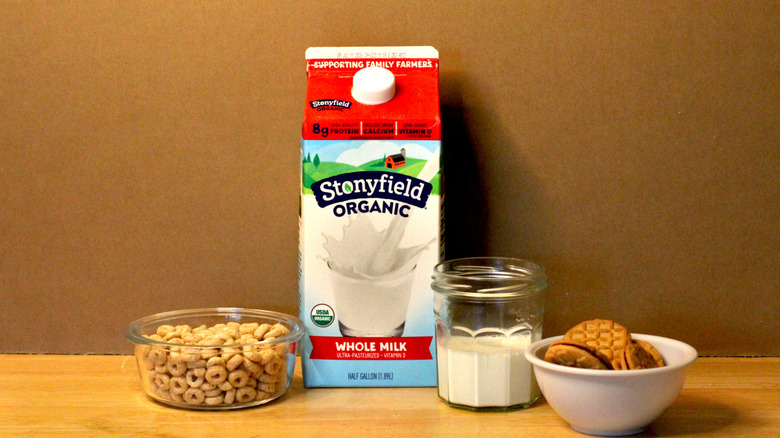 A Stonyfield brand carton of organic whole milk beside a glass of milk, a bowl of cereal, and some cookies
