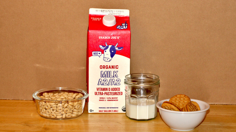 A Trader Joe's brand carton of organic A2 whole milk beside a glass of milk, a bowl of cereal, and some cookies
