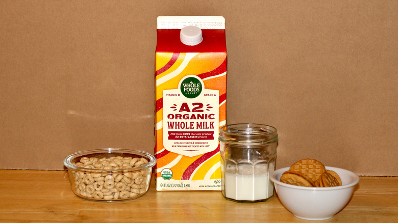 A Whole Foods brand carton of organic A2 whole milk beside a glass of milk, a bowl of cereal, and some cookies