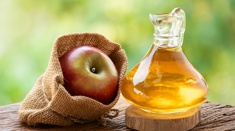 A jug of apple cider vinegar on a wooden table next to a burlap sack with an apple in it.