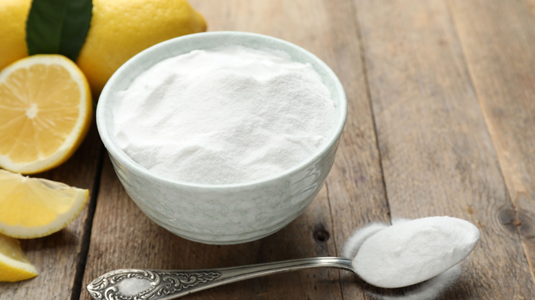 A bowl and spoon of baking soda on a table next to lemons