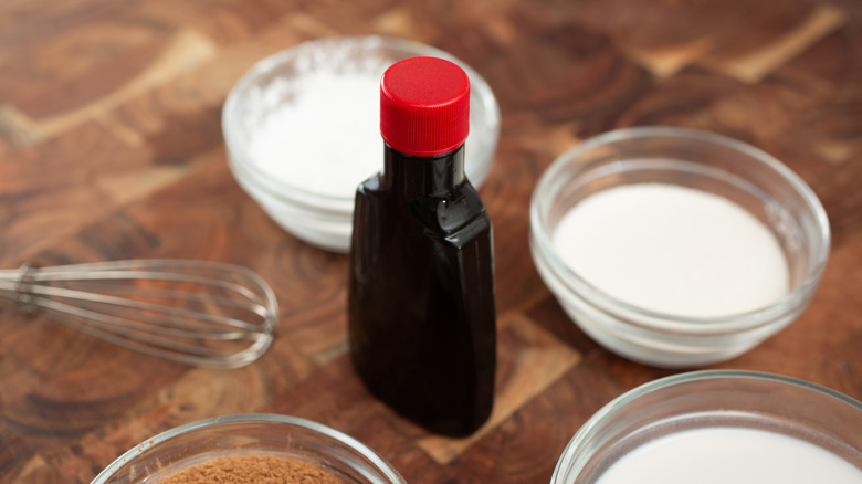A bottle of vanilla extract on a table surrounded by bowls of flour and sugar and a whisk