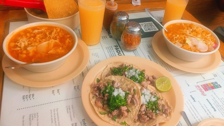 Bowls of pozole, a plate of tacos, chips, and orange-colored drinks on a table at La Casa de Toño