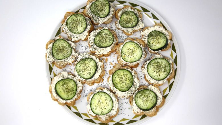 An overhead shot of a plate of open-faced cucumber sandwiches topped with dill on a white background