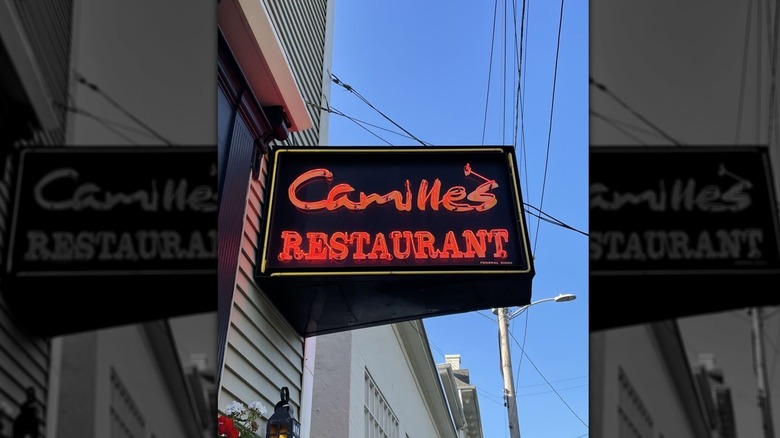 The exterior sign for Camille's at dusk attached to a gray-sided building; "Camille's Restaurant" is in red neon on a black background; electric wires criss-cross in the sky