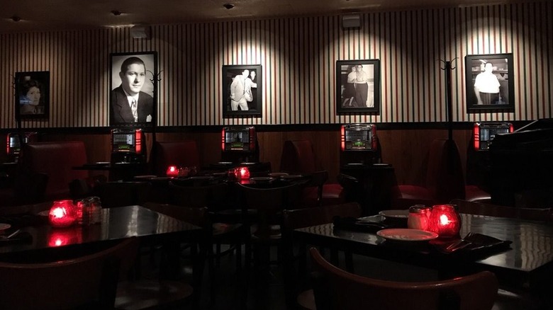 A dimly lit dining room at Campisi's with red votive candles on dark wood four-top tables and booths running along a back wall; black and white photos hang on a backdrop of white, red, and green pinstripe wallpaper