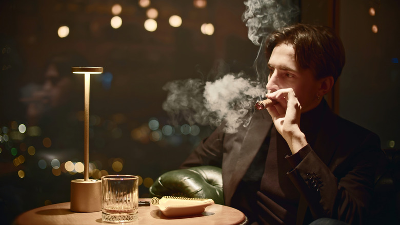 A man in a dark suit sitting at a restaurant table smoking a cigar; he is looking off to the side with a serious look on his face