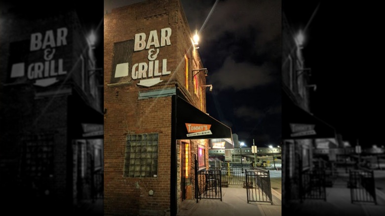The exterior of Tommy's from the side at night: Two-story brick building with a glass brick window on the first floor; "Bar & Grill" is painted in white with an arrow pointing down is painted on the second story; black awning on the front of the building over the entrance