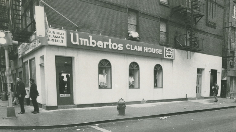 A black-and-white vintage photo of Umberto's Clam House on a corner of a city street; white stucco facade wih a door on the left and  three windows with rounded tops line the right side; sign reads "Umbertos Clam House" with a sign on the left that reads "scungilli, calamari, mussels" and a Pepsi logo sign on the right