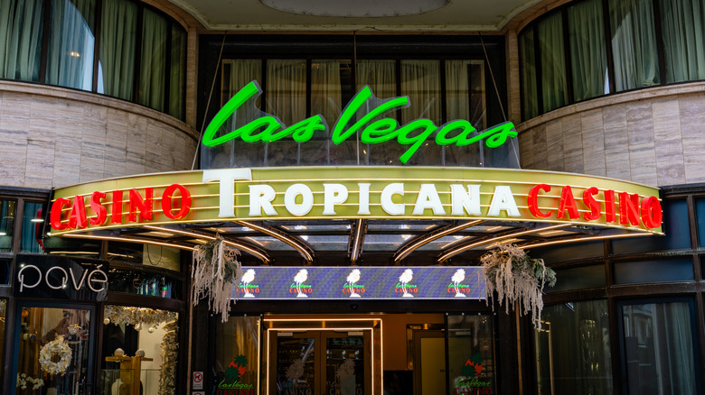 An exterior sign on the Tropicana Las Vegas casino at dusk; "Tropicana" is lit in white and centered over an entrance door with "casino" on each side lit in red; "Las Vegas" appears above it lit in green