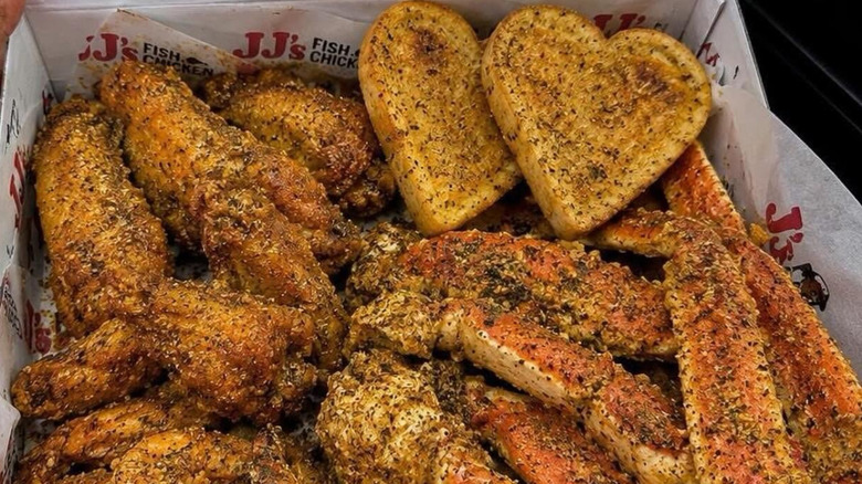 A JJ's Fish & Chip box filled with chicken wings, fried fish, crab legs and heart-shaped garlic bread.