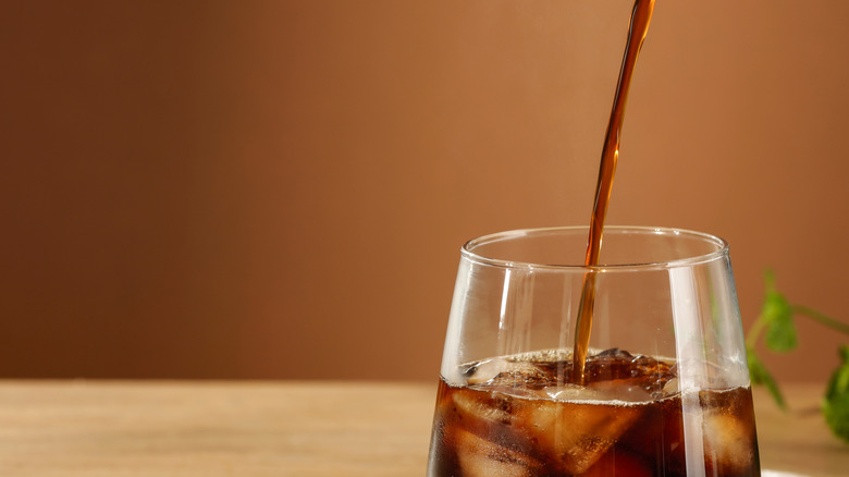 Cold brew coffee being poured into a glass filled with ice against a warm brown background.