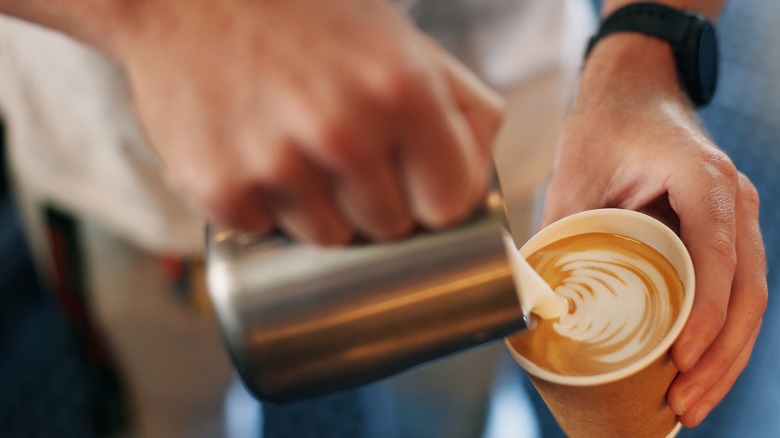 A barista pouring steamed milk into a paper cup, creating latte art.