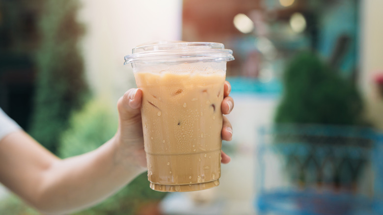 An iced latte in a plastic cup being hand by someone outdoors.