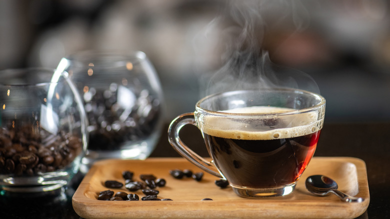 A glass cup of hot black coffee on a wooden tray with coffee beans scattered around, steam rising from the surface.