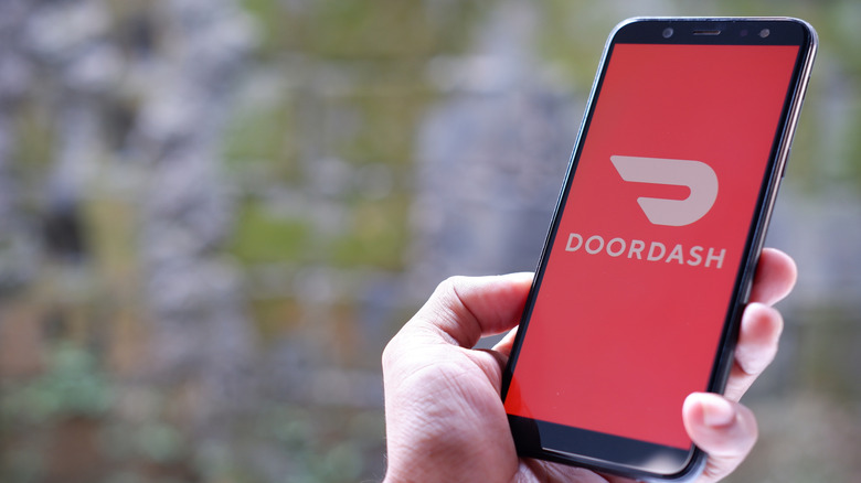 A hand holding a phone with the DoorDash logo on the screen against a blurred background