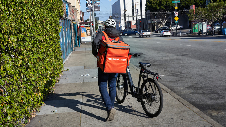 The back of a DoorDash delivery driver walking a bicycle along a city street