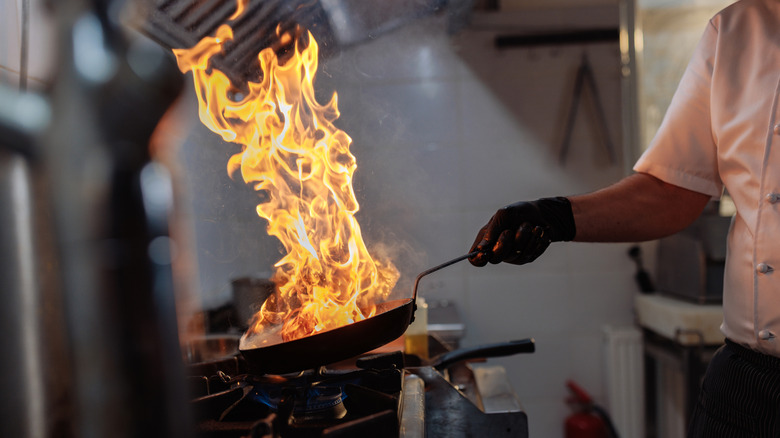 A chef holds a skillet over a stove in a professional kitchen with a large flame coming out of the pan