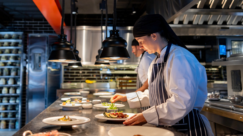 A chef in a striped apron plates food on a metal counter in a professional kitchen