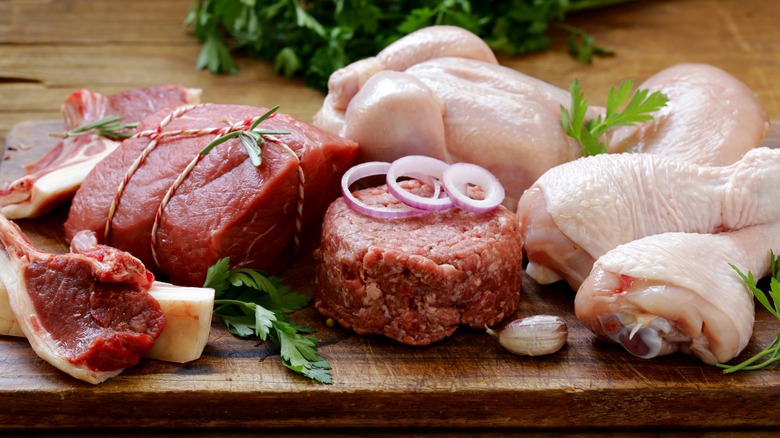 A selection of raw meats on a wooden cutting board among raw herbs and onions