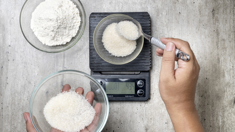 A person measures sugar from a bowl to a bowl sitting on a digital kitchen scale