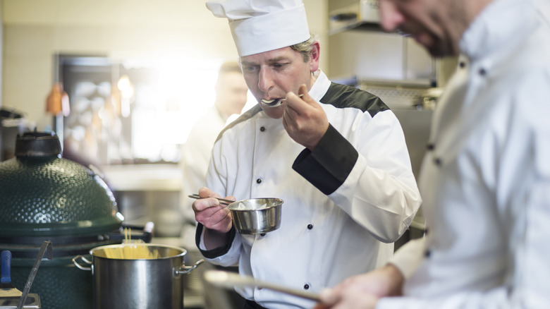 Two chefs in white coats and hats taste a sauce from a pot