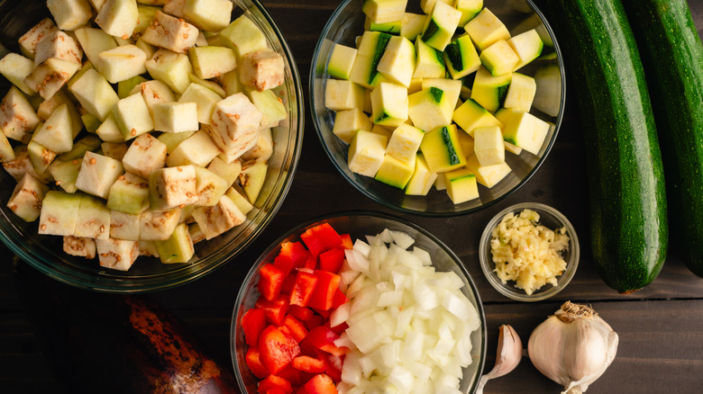 Bowls of cubed eggplant, zucchini, chopped bell pepper and onion, and minced garlic on a wooden counter