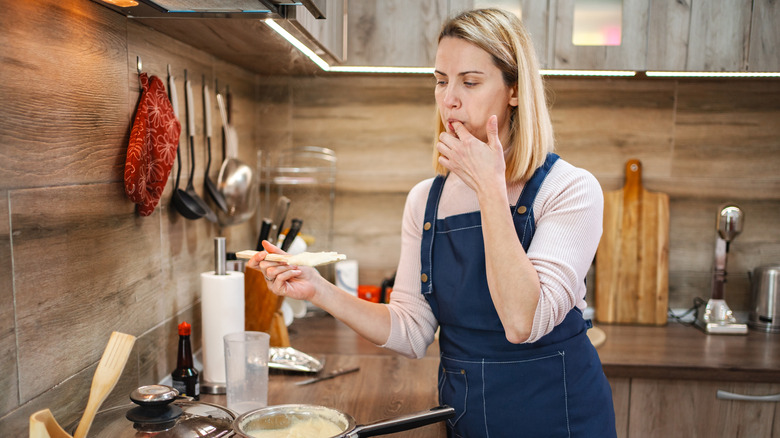 A female chef in a blue apron licks her finger after dipping it in a white sauce on the stove