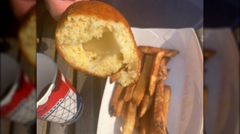 Someone holding a half-eaten cheese-on-a-stick above an order of fries and a drink