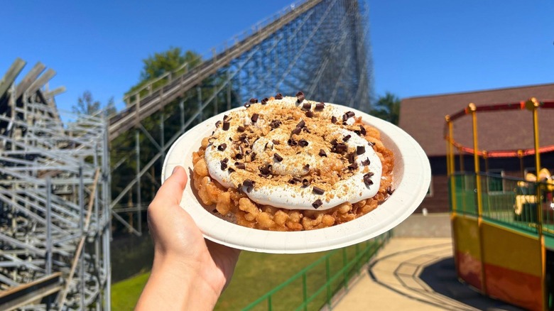 Someone holding a funnel cake in the air next to a rollercoaster