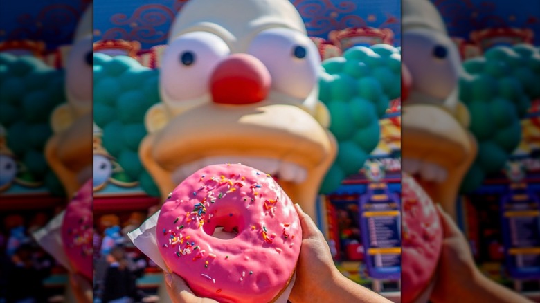 Someone holding up a pink donut in front of a clown-shaped structure
