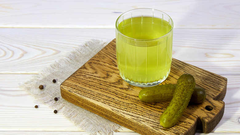 A glass of pickle juice with whole pickles on a wood board