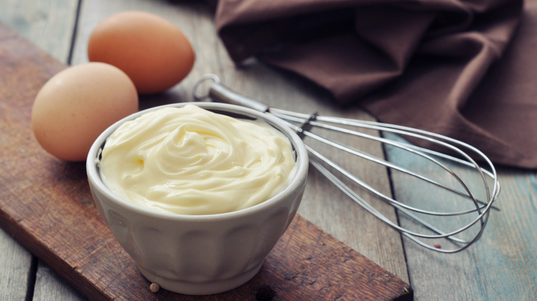 A ramekin of mayonnaise, eggs, and a whisk on a wood table
