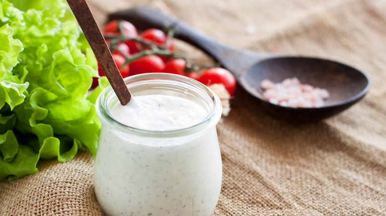A jar of white salad dressing next to lettuce and tomatoes on a burlap background