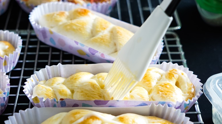 A baking brush brushing mayonnaise onto small loaves of braided bread in colorful liners