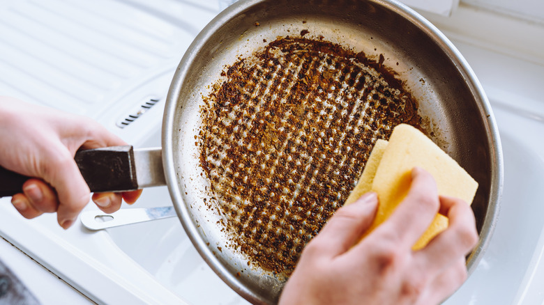 A woman's hand holding a dirty pan over the sink and scrubbing it with a sponge