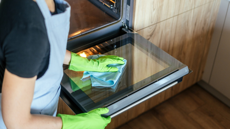 A woman in kitchen gloves is focused on cleaning the oven door with a cloth.