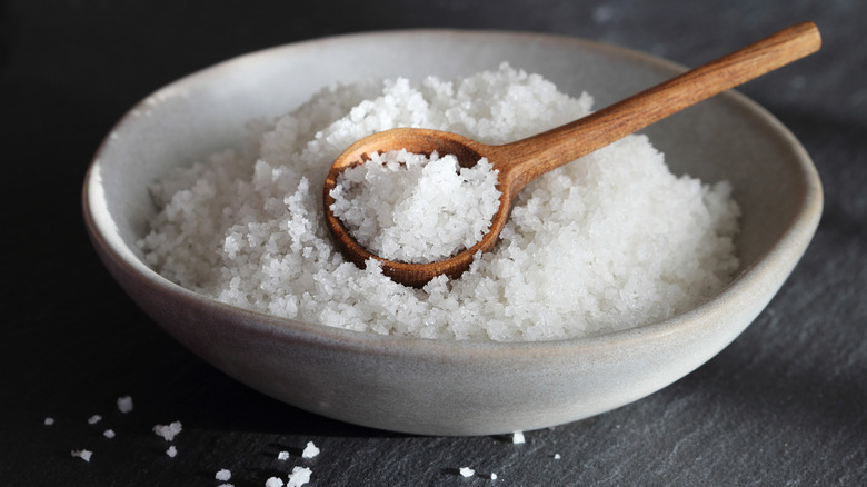 Big granules of sea salt in a ceramic grey bowl with wooden spoon on a black slate.