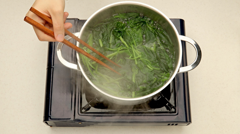 A picture of a hand blanching spinach in hot water with chopsticks.