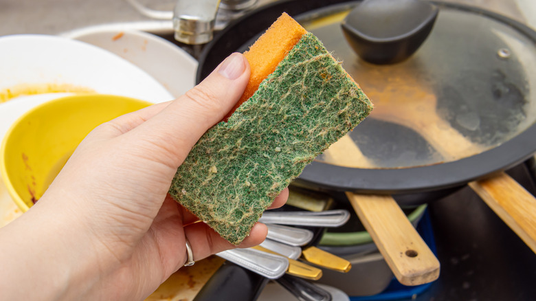 Woman holds orange sponge for washing dishes in front of sink filled with dirty pans, plates, and utensils