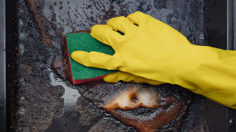 A yellow rubber gloved hand cleaning a dirty baking sheet with a sponge