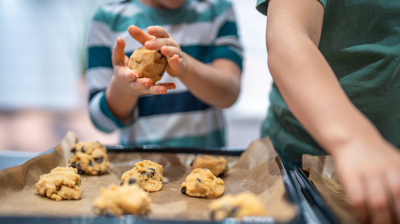 A baking sheet covered in parchment paper with cookie dough as two children shape the dough