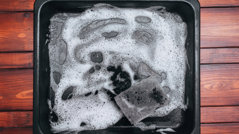 A black baking sheet with soap bubbles and scrubbing pad on wooden surface