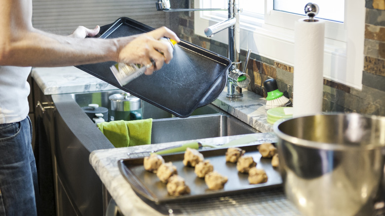 A man spraying oil on a baking sheet over sink next to another baking sheet with balls of cookie dough in kitchen