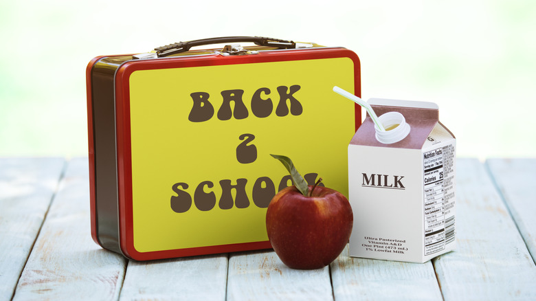 A metal lunchbox bearing the words "back 2 school" shown with a red apple and a half-pint carton of milk with a straw