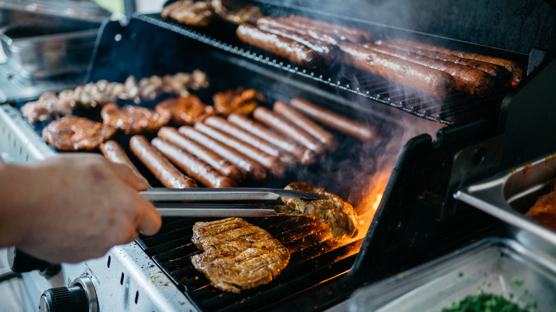 Someone flipping a piece of chicken over on a flaming grill with hot dogs and other meat in the background.