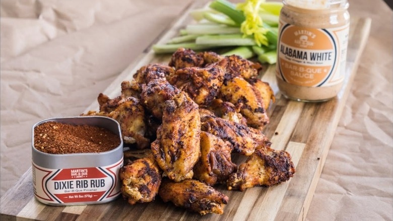 pile of martin's bbq joint chicken wings on a wooden chopping board next to martin's signature white sauce and dry rub and a pile of celery sticks